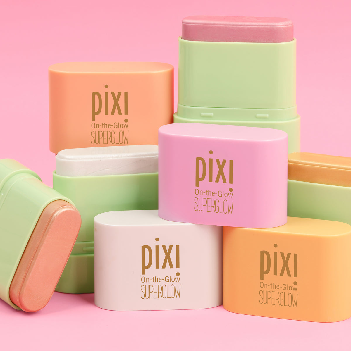 A group of Pixi On-the-Glow SuperGlow stick products in pastel shades of pink, orange, yellow, and white, arranged on a solid pink background -