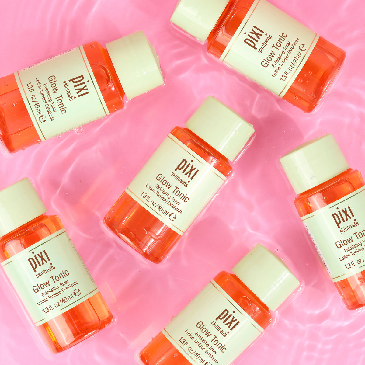 Six small Pixi Glow Tonic bottles arranged on a pink water-textured background. The bottles are translucent orange with cream-colored caps and labels, and the brand name Pixi is clearly visible.