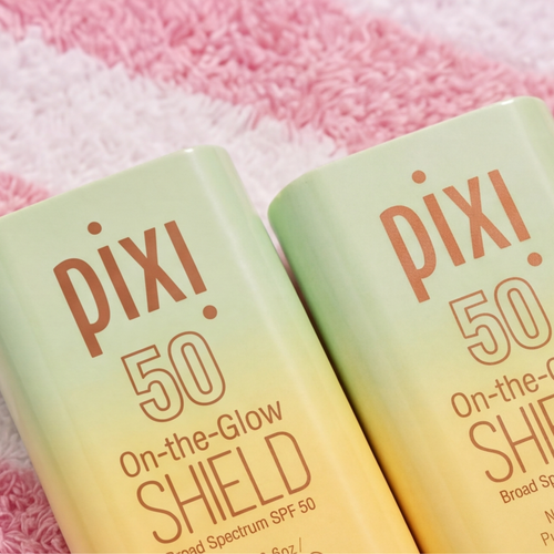Two Pixi On-the-Glow Shield Broad Spectrum SPF 50 sunscreen bottles with a light green to pale yellow gradient, resting on a pink textured towel. view 2