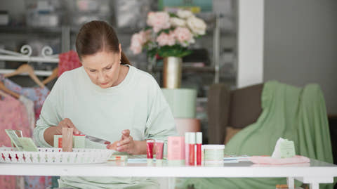 A woman in a light sage green sweatshirt sits at a white table, applying makeup from a small pink tube to her hand. Several cosmetic products in pink, red, and white packaging are arranged on the table in a softly lit room with a blurred background of clothing racks and flowers.