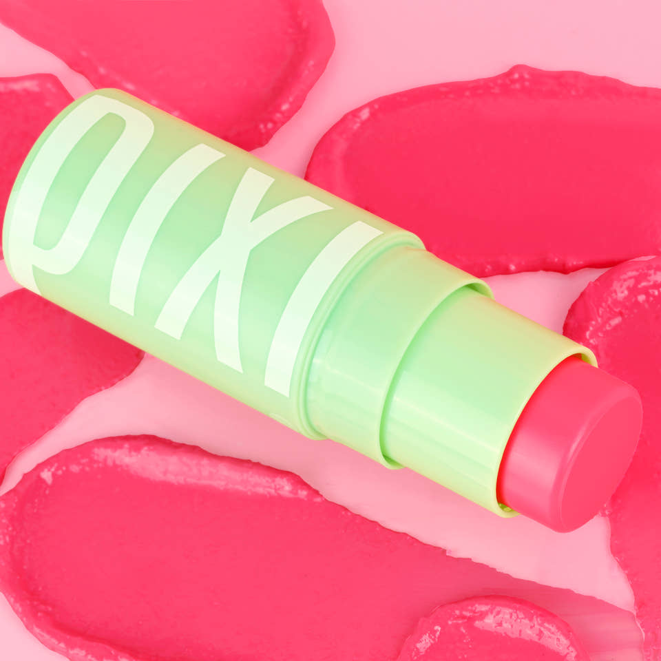 A close-up of a Pixi +Hydra LipTreats tube lying diagonally on a pink background with pink lip-shaped decorative elements. The tube is light green with large white Pixi lettering, and the balm stick itself is a bright pink. view 1