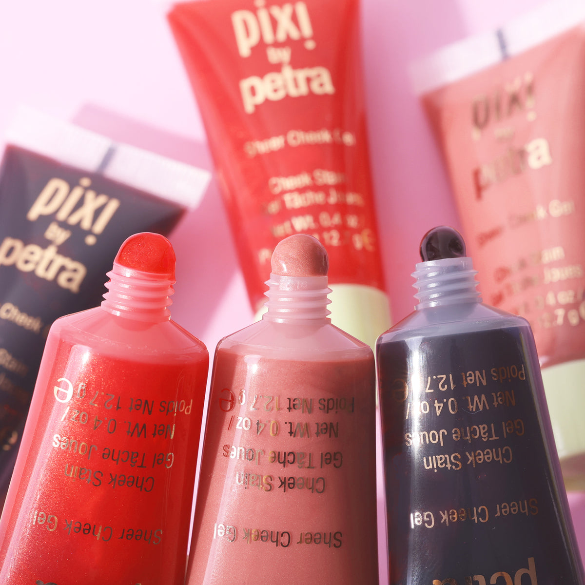 Three open Sheer Cheek Gel tubes of Pixi by Petra cheek stain in shades of coral red, nude pink, and dark charcoal, arranged in a close-up studio shot against a soft pink background.