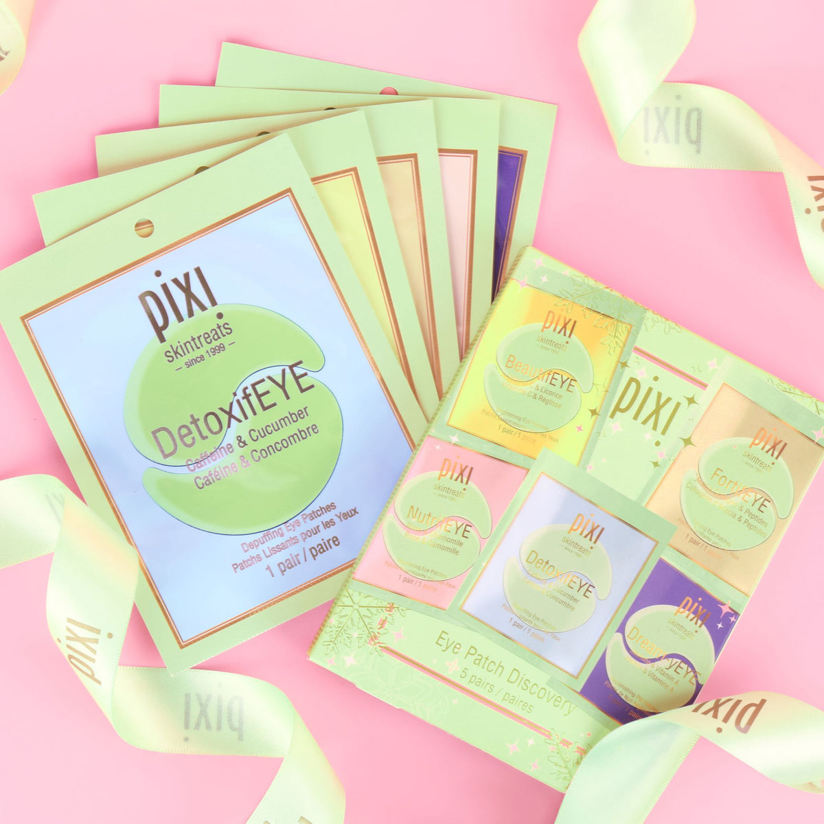 A flat-lay of Pixi eye patch packets on a pastel pink background with decorative white ribbons. The main packet is light green with a blue center panel, and a second packet shows multiple individual eye patch discs in pastel yellow, pink, green, and purple.