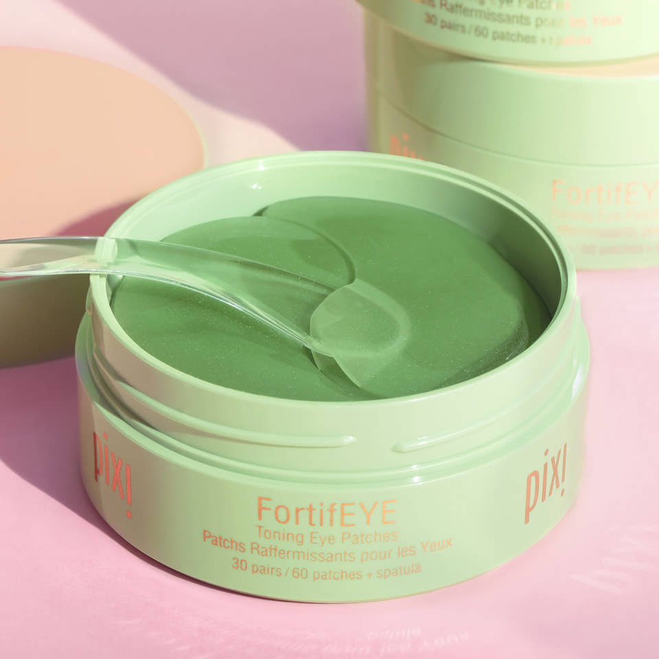 An open round jar of Pixi FortifEYE toning eye patches in a mint green shade, with a clear plastic spatula resting inside. The background is a soft pink with a second closed jar blurred in the background. view 1